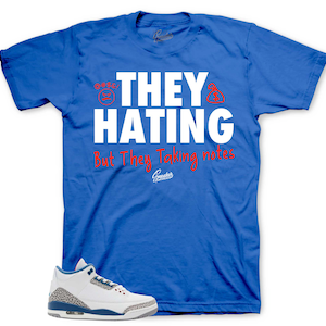 May include: A royal blue short-sleeve t-shirt featuring the white text "THEY HATING" with red money bag icons, and the red text "But They Taking notes" below. A white, grey, and blue athletic sneaker is positioned at the bottom left of the shirt.