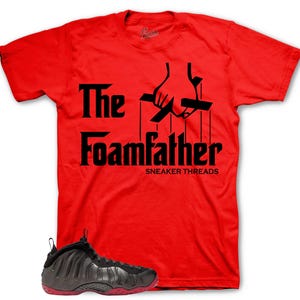 May include: A red t-shirt with the black text "The Foamfather" and a puppet hand design. A black and grey trainer with a red sole is in the foreground. The shirt is a short-sleeved crew neck.