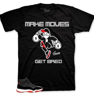 May include: Black t-shirt with white text that reads "MAKE MOVES GET BREAD". A cartoon character wearing a red hat and mask is holding money bags. The character is wearing red shoes. A pair of black and red sneakers are in the bottom left corner of the image.