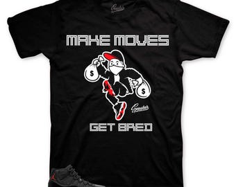 May include: Black t-shirt with white text that reads "MAKE MOVES GET BREAD". A cartoon character wearing a red hat and mask is holding money bags. The character is wearing red shoes. A pair of black and red sneakers are in the bottom left corner of the image.