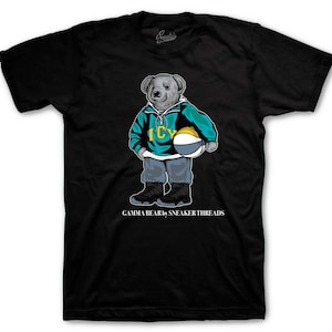 May include: Black t-shirt featuring a cartoon bear wearing a teal and yellow jacket, gray pants, and black sneakers. The bear is holding a basketball. The text "ICY" is on the jacket, and "GAMMA BEAR by SNEAKER THREADS" is at the bottom.