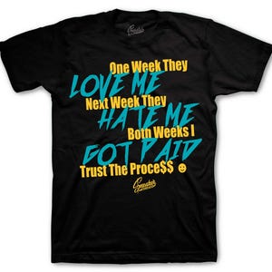 May include: Black t-shirt with the text "One Week They Love Me, Next Week They Hate Me, Both Weeks I Got Paid, Trust The Process$" in teal and yellow. The shirt has a crew neck.