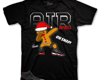 May include: Black t-shirt with a gingerbread man wearing a Santa hat and doing the dab. The text "AIR BRED" and "OH SNAP!" is printed on the shirt.
