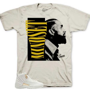 May include: Cream-colored t-shirt featuring a black and yellow graphic of a man's profile and the word "Hustle". A pair of white and gold sneakers is in the foreground. The shirt has a crew neck and short sleeves.