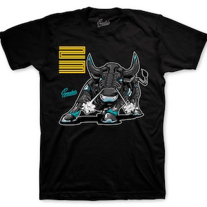 May include: Black t-shirt featuring a stylized illustration of a bull with teal and black accents. The bull has a robotic design with steam coming from its nostrils. The shirt also has a yellow and black design and the word "Sneakers" in teal.