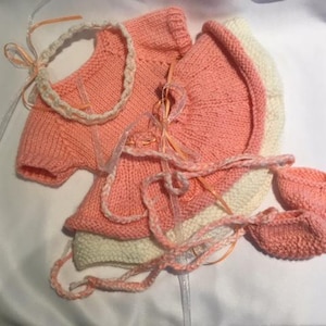 May include: A pink knitted baby outfit set with a matching headband, dress, and booties. The set is made of soft yarn and is perfect for a newborn baby.