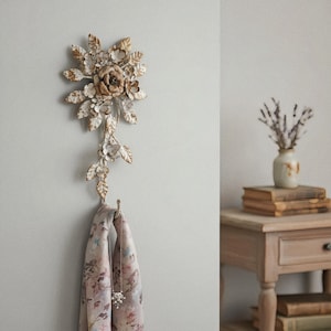 May include: A decorative metal wall hook in the shape of a floral arrangement. The hook is painted in shades of white and gold, with a scarf and necklace hanging from it. The background is a light gray wall.