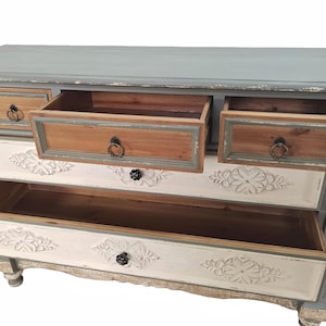May include: A distressed-finish chest of drawers with a light blue top and gray frame. The drawers are a mix of natural wood, white, and gray, with carved floral details. Some drawers are open, revealing the interior.