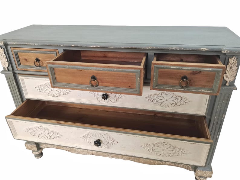 May include: A distressed-finish chest of drawers with a light blue top and gray frame. The drawers are a mix of natural wood, white, and gray, with carved floral details. Some drawers are open, revealing the interior.