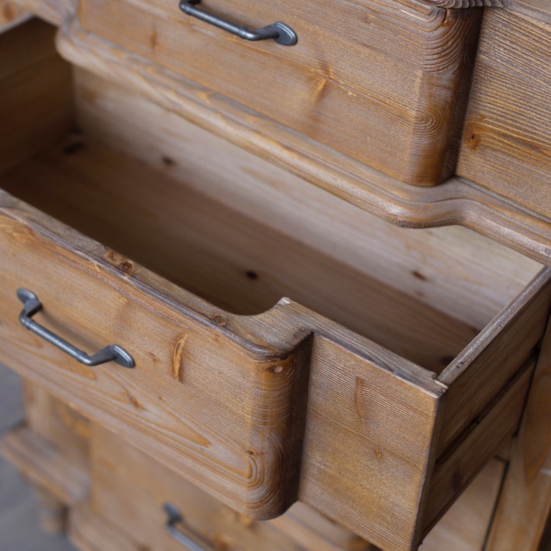 May include: A wooden chest of drawers with multiple drawers. The top drawer is open, revealing the empty interior. The drawers have black metal handles.