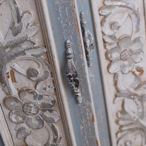 Op de afbeelding: Close-up van een verweerde, antieke kastdeur met sierlijke bloemsnijwerken. De deur heeft een lichtblauw en wit kleurenschema met zilverkleurige hardware. De snijwerken zijn gedetailleerd en voegen een vintage esthetiek toe.