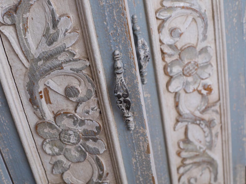 Op de afbeelding: Close-up van een verweerde, antieke kastdeur met sierlijke bloemsnijwerken. De deur heeft een lichtblauw en wit kleurenschema met zilverkleurige hardware. De snijwerken zijn gedetailleerd en voegen een vintage esthetiek toe.