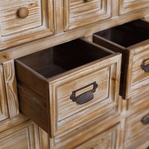 May include: A wooden cabinet with multiple drawers, some open. The cabinet has a light brown, distressed finish. The open drawers reveal a dark interior. Each drawer has a metal handle. The cabinet is made of wood.