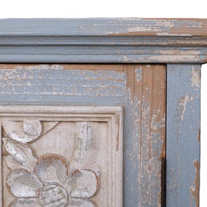 Op de afbeelding: Close-up van een hoek van een verweerde kast. De kast heeft een lichtblauw en beige kleurenschema met een gebeeldhouwd bloemmotief. De verf heeft een verweerde, afbladderende uitstraling, wat bijdraagt aan de vintage esthetiek.