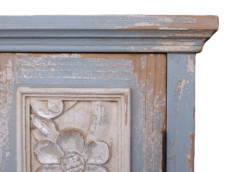 Op de afbeelding: Close-up van een hoek van een verweerde kast. De kast heeft een lichtblauw en beige kleurenschema met een gebeeldhouwd bloemmotief. De verf heeft een verweerde, afbladderende uitstraling, wat bijdraagt aan de vintage esthetiek.