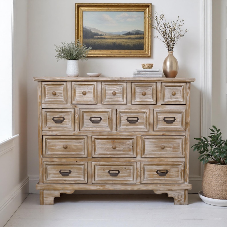 May include: A light-toned wooden chest of drawers with numerous square drawers, each with a small metal handle. The chest is set against a white wall, with a framed landscape painting and decorative items on top.