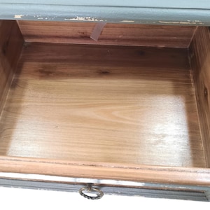 May include: An open wooden drawer with a light brown interior and a gray-blue painted frame. The drawer has a metal ring pull. The wood grain is visible on the drawer's interior and frame.