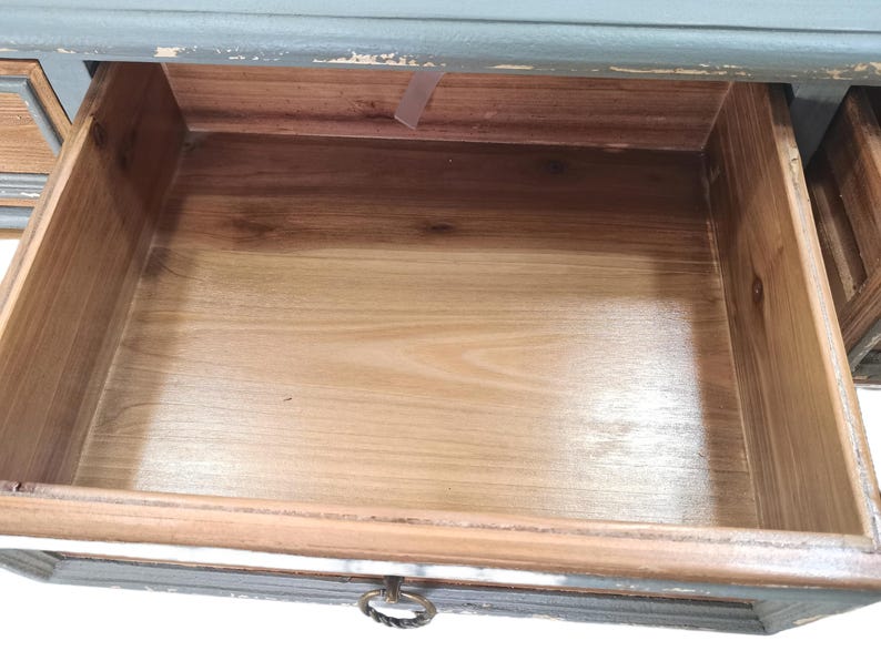 May include: An open wooden drawer with a light brown interior and a gray-blue painted frame. The drawer has a metal ring pull. The wood grain is visible on the drawer's interior and frame.