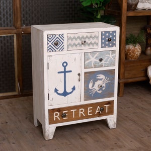 Puede incluir: Un mueble de madera blanco con acabado envejecido, con seis cajones y una puerta. Decorado con diseños de temática náutica, incluyendo un ancla, estrellas de mar y cangrejos. La palabra "RETREAT" está impresa en el cajón inferior. Mide unos 70 cm de alto.