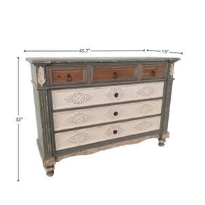 May include: A distressed-finish chest of drawers, 45.7" wide and 32" tall. The top drawers are brown, the middle drawers are white with floral carvings, and the frame is gray. The chest has decorative accents and black knobs.