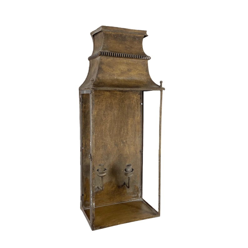 May include: A vintage-style wall lantern with a rectangular frame and a decorative top. The lantern is made of a bronze-colored metal and features two candle holders inside. The design is reminiscent of antique lighting fixtures.