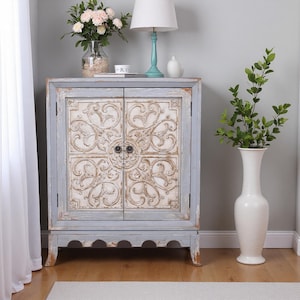 Puede incluir: Un mueble envejecido de dos puertas con un diseño tallado ornamentado en crema y blanco. El mueble tiene un marco y una base azul claro. Una lámpara blanca, un jarrón de flores y un jarrón blanco alto con vegetación están cerca.