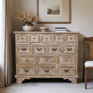 May include: A light-coloured wooden chest of drawers with multiple drawers and bronze-toned hardware. The chest has a rustic, weathered appearance and is topped with decorative items, including a vase with dried plants and framed artwork. A wooden chair is to the right.