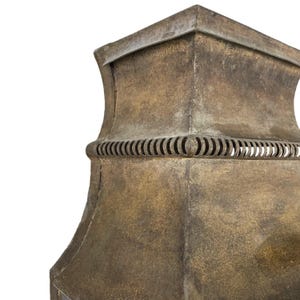 May include: Close-up of a weathered, bronze-colored metal lantern. The lantern has a square top and a decorative band of curved cutouts. The metal has a textured, aged appearance, suggesting it is an antique or vintage piece.