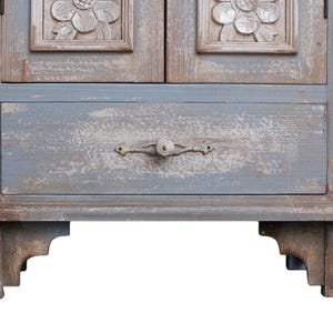 Op de afbeelding: Een verweerde, lichtblauwe houten kast met twee deuren en een lade. De deuren hebben gesneden bloemendetails. De lade heeft een decoratieve metalen handgreep. De kast staat op decoratieve poten en toont een rustieke, vintage esthetiek.