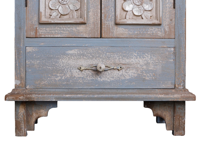 Op de afbeelding: Een verweerde, lichtblauwe houten kast met twee deuren en een lade. De deuren hebben gesneden bloemendetails. De lade heeft een decoratieve metalen handgreep. De kast staat op decoratieve poten en toont een rustieke, vintage esthetiek.