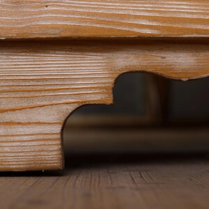 May include: Close-up of a wooden furniture corner joint. The wood has a warm, light brown tone with visible grain patterns. The design includes a decorative, curved cutout detail, adding visual interest to the structure.