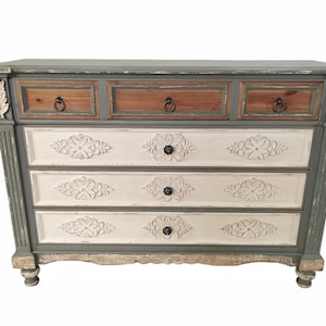 May include: A distressed-finish chest of drawers with three small top drawers and three larger drawers below. The top drawers are brown, the middle drawers are white with floral carvings, and the frame is gray. The chest has decorative accents and ring pulls.