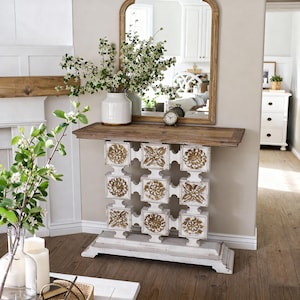 Puede incluir: Una consola blanca y marrón con un diseño floral decorativo. La mesa tiene una tapa de madera y una base blanca con detalles florales tallados. Un jarrón blanco con vegetación y un pequeño reloj encima.