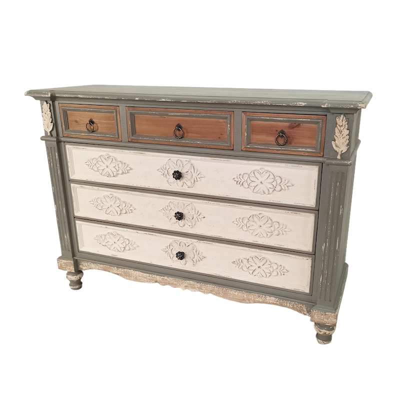 May include: A distressed-finish chest of drawers with a sage green and white color scheme. The top drawers are wood-toned, while the lower drawers are white with floral carvings. The chest has decorative accents and ring pulls.