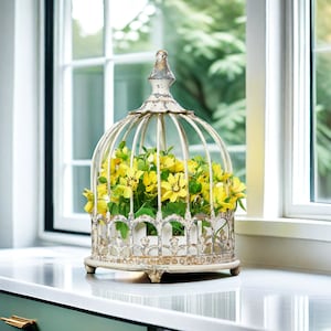 May include: A decorative, distressed white metal birdcage filled with yellow flowers and green leaves. The cage has a domed top and ornate detailing. It sits on a white countertop near a window, creating a charming, vintage-inspired display.