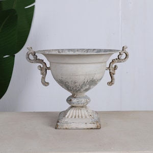 Pode incluir: Uma urna de metal branco envelhecido com alças ornamentadas. A urna tem uma base de pedestal e uma base quadrada. A tinta está lascada, dando-lhe um aspeto vintage. Adequada para uso interno ou externo.