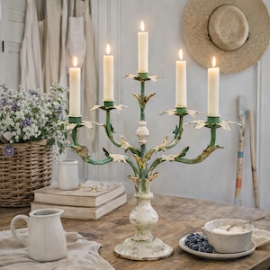 Könnte beinhalten: Ein Kerzenleuchter im Vintage-Stil mit fünf brennenden Kerzen. Der Kerzenleuchter hat eine verwitterte grün-cremefarbene Oberfläche mit Blättern. Die Szene umfasst einen Blumenkorb und einen weißen Krug.