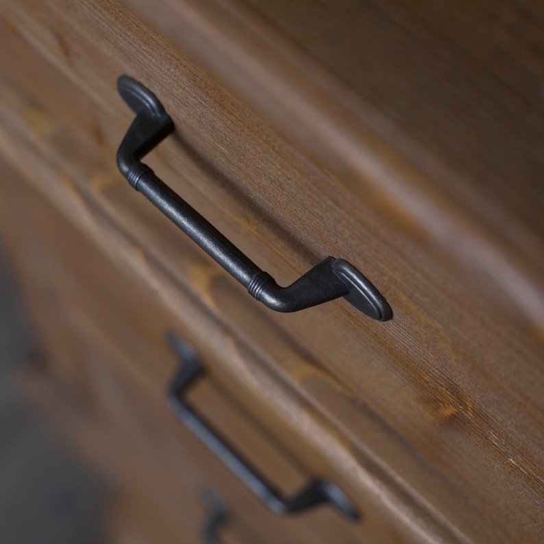 May include: Close-up of a wooden chest of drawers with a black metal handle. The handle is a simple, rectangular design with rounded edges.