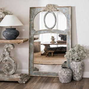 May include: A large, rectangular mirror with a distressed light blue and grey frame. An oval mirror is centered above the main mirror. The mirror is placed next to a distressed wooden table with a lamp and decorative vases.
