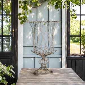 May include: A decorative metal planter with a lattice design. The planter has a wide, flared top and a pedestal base. It is made of a light-coloured metal and sits on a wooden table. The background includes a blue wall and a dark door.