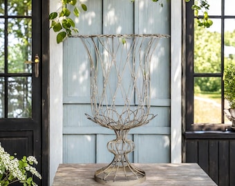 Op de afbeelding: Een hoge, decoratieve metalen plantenbak met een traliewerkontwerp. De plantenbak heeft een brede, uitlopende bovenkant en een voetstuk. Het is gemaakt van een lichtgekleurd metaal en staat op een houten tafel. De achtergrond omvat een blauwe muur en een donkere deur.