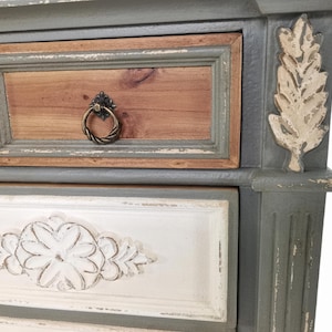 May include: Close-up of a distressed, painted wooden dresser. The drawers feature a mix of colors, including gray, brown, and white. Decorative leaf accents and ornate drawer pulls add to the vintage aesthetic.