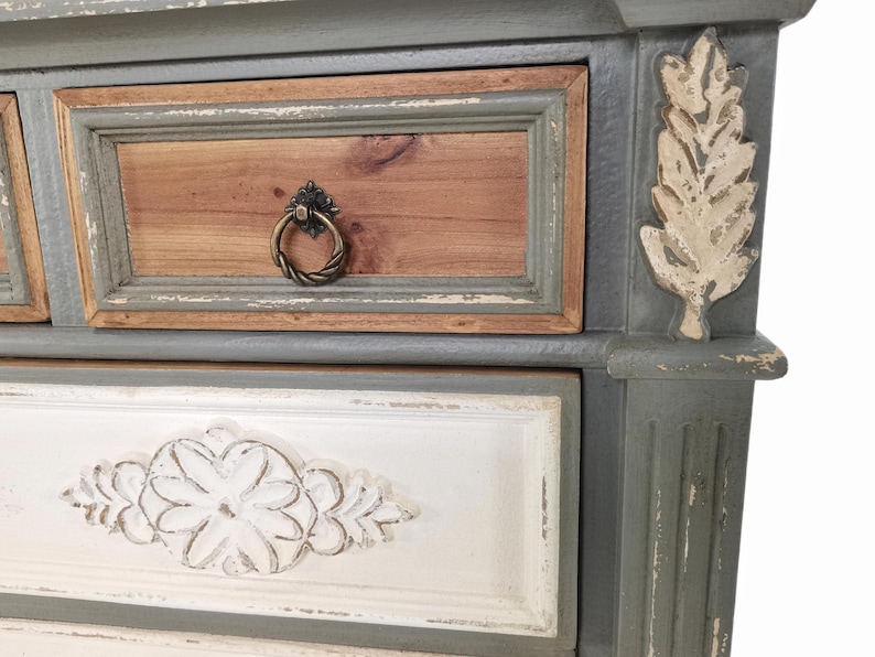 May include: Close-up of a distressed, painted wooden dresser. The drawers feature a mix of colors, including gray, brown, and white. Decorative leaf accents and ornate drawer pulls add to the vintage aesthetic.