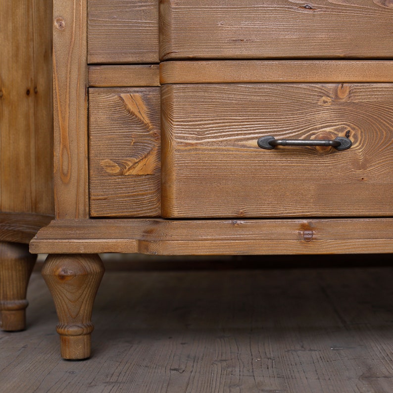 May include: A wooden chest of drawers with four drawers and turned legs. The dresser is made of light brown wood with a natural finish. The drawers have black metal handles.