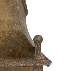 May include: Close-up of a weathered, antique-style metal lantern. The lantern has a tarnished, golden-brown finish with intricate details. A small, rounded knob sits atop the lantern's roof, and a small water droplet is visible.