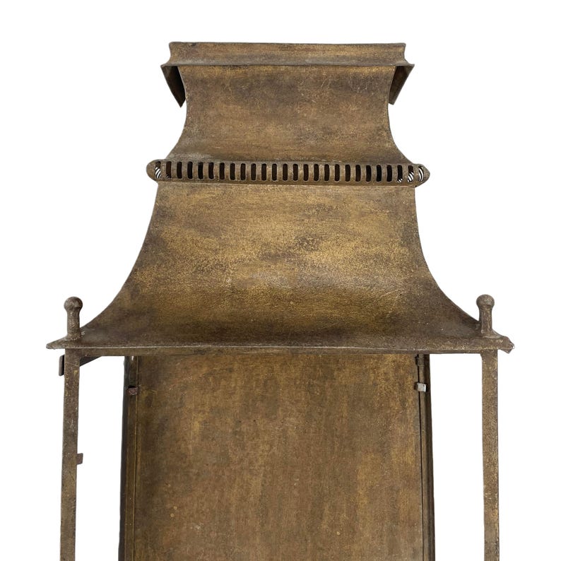 May include: A close-up of a bronze-colored lantern with a pagoda-style top. The lantern has a rectangular body and a decorative, curved roof. The metal has a textured, aged appearance, with small decorative finials on the corners.