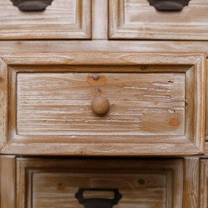 May include: Close-up of a wooden cabinet with multiple drawers. The drawers are made of light-coloured wood with a distressed finish, featuring a round wooden knob. The cabinet has a rustic, vintage aesthetic.