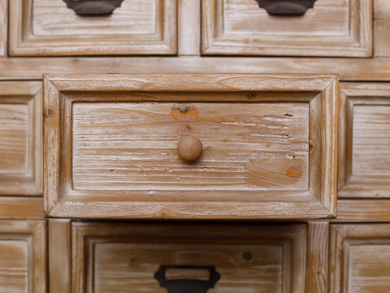 May include: Close-up of a wooden cabinet with multiple drawers. The drawers are made of light-coloured wood with a distressed finish, featuring a round wooden knob. The cabinet has a rustic, vintage aesthetic.
