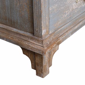 Op de afbeelding: Close-up van een hoek van een verweerde houten kast. De kast heeft een lichtblauwe en bruine afwerking met zichtbare houtnerf en decoratieve lijstwerk. De hoek heeft een gedetailleerde, gesneden poot.