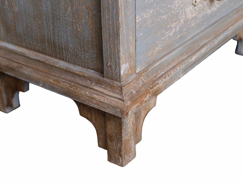 Op de afbeelding: Close-up van een hoek van een verweerde houten kast. De kast heeft een lichtblauwe en bruine afwerking met zichtbare houtnerf en decoratieve lijstwerk. De hoek heeft een gedetailleerde, gesneden poot.